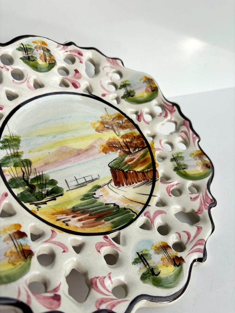 May include: White ceramic plate with a hand-painted landscape scene of trees, hills, and a body of water. The plate has a scalloped edge and a delicate lace-like design around the rim.