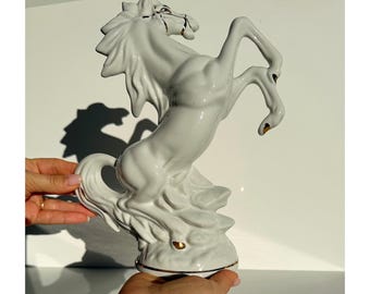 May include: A white ceramic horse statue rearing up on its hind legs. The horse has a flowing mane and tail, with gold accents on the hooves and bridle. The statue is a decorative item.