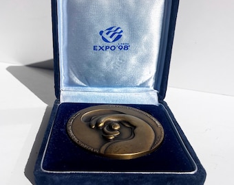 Könnte beinhalten: Bronzefarbene Medaille in einer blauen Samtbox. Die Medaille zeigt eine Hand, die einen Globus hält. Auf der Box steht "EXPO'98" und ein Logo.