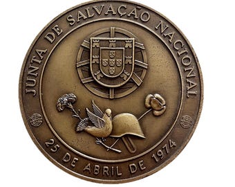 May include: A bronze-colored commemorative coin with the text "JUNTA DE SALVAÇÃO NACIONAL" and "25 DE ABRIL DE 1974". The coin features a coat of arms, a dove, a helmet, and flowers. The coin has a circular shape.