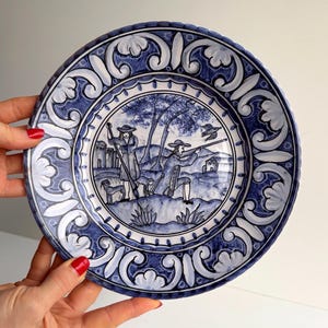Könnte beinhalten: Blaue und weiße Keramikplatte mit handgemalter Jagdszene. Die Platte hat einen dekorativen Rand mit floralen und Rankenmustern. Der Durchmesser der Platte beträgt ca. 25 cm. Die Platte wird von einer Person gehalten.