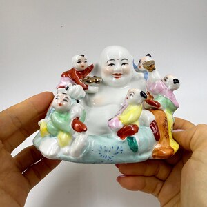 May include: A white ceramic figurine of a smiling Buddha surrounded by small figures in colorful clothing. The Buddha is holding a gold object. The base is light blue with a floral pattern. The figurine is held by a person's hands.
