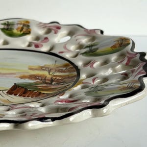 May include: A white ceramic plate with a scalloped edge and a black rim. The plate has a hand-painted design of a landscape with trees and a fence. The plate also has a pattern of small holes around the edge.