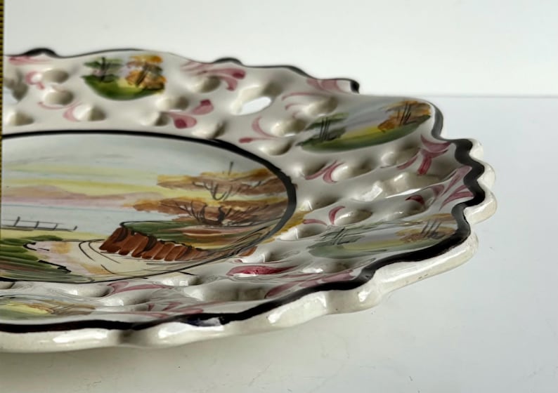 May include: A white ceramic plate with a scalloped edge and a black rim. The plate has a hand-painted design of a landscape with trees and a fence. The plate also has a pattern of small holes around the edge.
