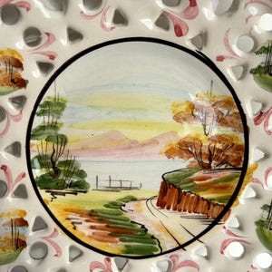 May include: A white ceramic plate with a cut-out design around the edge. The center of the plate features a hand-painted landscape scene with a lake, trees, and a path leading to a small wooden bridge. The scene is painted in shades of green, brown, yellow, and pink.