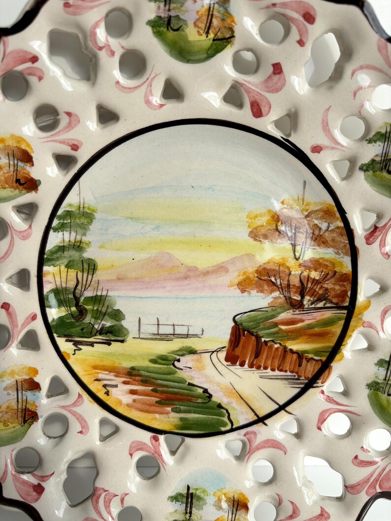 May include: A white ceramic plate with a cut-out design around the edge. The center of the plate features a hand-painted landscape scene with a lake, trees, and a path leading to a small wooden bridge. The scene is painted in shades of green, brown, yellow, and pink.