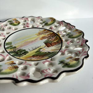 May include: A white ceramic plate with a scalloped edge and a lace-like design. The plate features a hand-painted scene of a landscape with trees, a body of water, and a path.