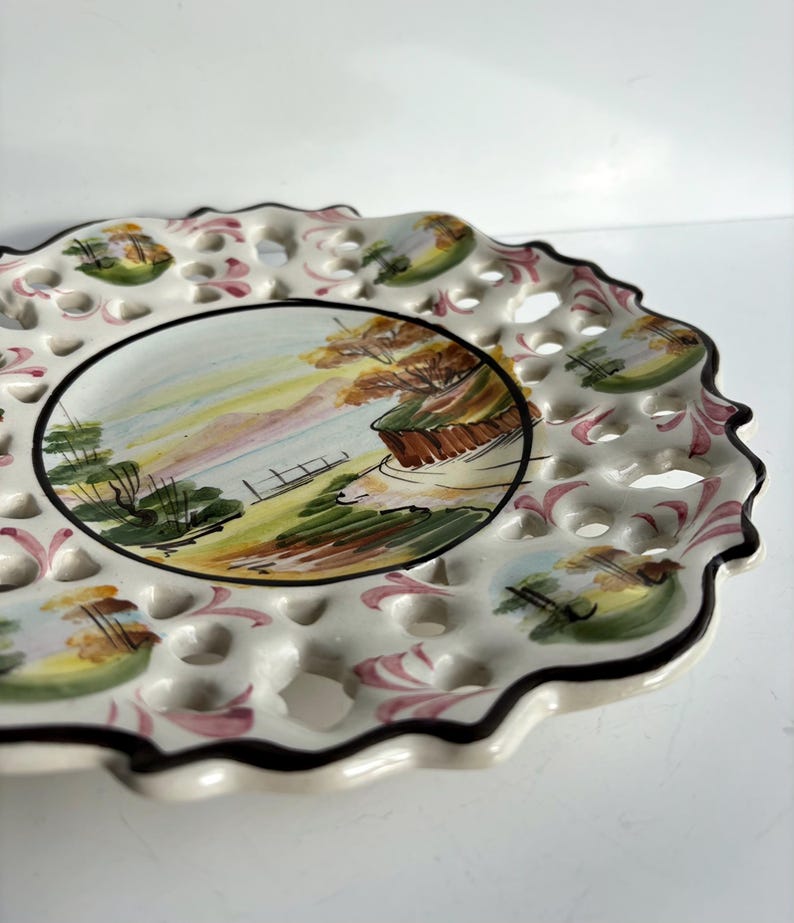 May include: A white ceramic plate with a scalloped edge and a lace-like design. The plate features a hand-painted scene of a landscape with trees, a body of water, and a path.