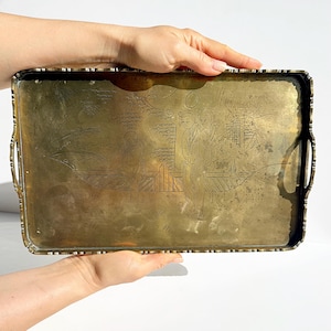 May include: Rectangular brass tray with etched Asian-inspired figures and landscape scenes. The tray has handles on the sides and a decorative edge. The brass has a warm, aged patina.