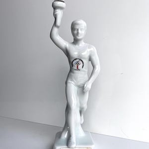 May include: A white ceramic figurine of a male athlete holding a torch aloft. The figurine has a logo on the chest and the text "AMSTERDAM HEEFT OLYMPISCH VUUR" on the base. The torch has a brown wick.