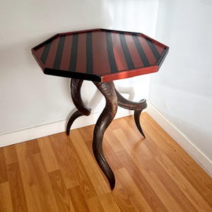 May include: A hexagonal side table featuring a flat top with alternating black and dark red stripes. The table is supported by three curved, dark brown, textured horn-like legs that extend outward and downward to the floor in a tripod configuration.