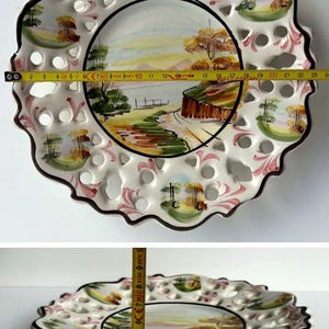 May include: A white ceramic plate with a scalloped edge and a cut-out design. The plate features a hand-painted landscape scene with trees, a river, and a bridge. The plate is approximately 23 inches in diameter.