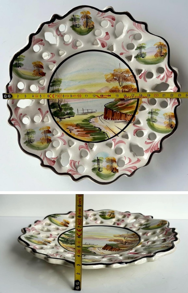 May include: A white ceramic plate with a scalloped edge and a cut-out design. The plate features a hand-painted landscape scene with trees, a river, and a bridge. The plate is approximately 23 inches in diameter.