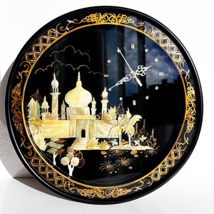 May include: A round black clock with a gold frame and a white and gold inlay of a mosque with palm trees. The clock face is behind a clear glass cover.