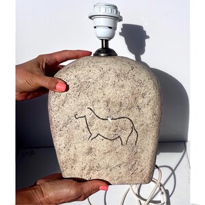May include: A beige ceramic table lamp with a textured surface and a black outline drawing of an animal. The lamp has a white bulb socket and a black metal base. The lamp is held by a person.