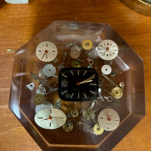 Puede incluir: Un pisapapeles octogonal transparente lleno de varias piezas de reloj y una esfera de reloj negra. La esfera del reloj tiene agujas blancas y un borde blanco.