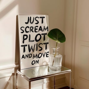 Puede incluir: Una impresión enmarcada en blanco con texto negro que dice "Just Scream Plot Twist And Move On". La impresión está sobre una mesa de vidrio con un jarrón de vidrio transparente que contiene una planta verde.