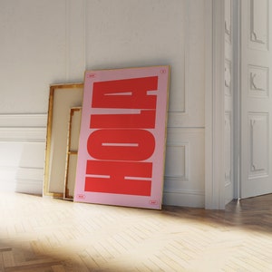 Op de afbeelding: Een roze en rode poster met het woord "HOLA" in grote, vetgedrukte, rode letters. De poster leunt tegen een witte muur, naast een houten frame.