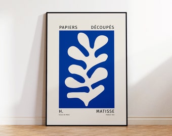 May include: A framed art print featuring a white abstract leaf design on a vibrant blue background. The print includes the text "PAPIERS DÉCOUPÉS" and "MATISSE FRANCE 1955". The artwork is displayed on a wooden floor.