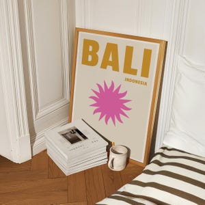 Peut inclure: Une affiche encadrée avec un cadre doré et un fond beige. L'affiche présente le mot "BALI" en grandes lettres jaunes et audacieuses et le mot "INDONESIA" en lettres noires plus petites. Un soleil rose à 12 rayons est centré sur l'affiche.