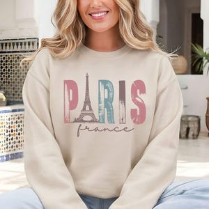 May include: Beige sweatshirt with the word "PARIS" in pink, blue, and pink, with an Eiffel Tower graphic. The word "france" is written below in a cursive font. The sweatshirt is a neutral color.
