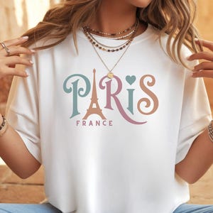 May include: White t-shirt with the word "Paris" in teal, pink, and brown script, featuring an Eiffel Tower graphic. The word "France" is printed below the word "Paris".