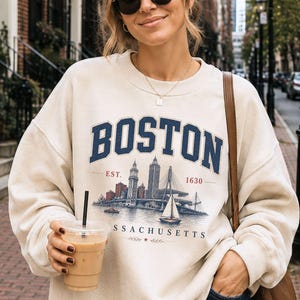 May include: A beige crewneck sweatshirt featuring a navy blue graphic print that reads BOSTON EST. 1630 MASSACHUSETTS. The design includes a detailed illustration of the Boston skyline, a bridge, and a sailboat on the water in shades of blue and red.