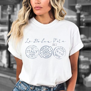 May include: White t-shirt with the text "La Dolce Vita 186.1" and three blue stamp-like images that say "Italy" with details like "Rome" and "Made in Italy".