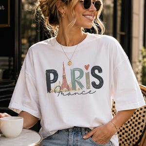 May include: A white short-sleeved t-shirt featuring a graphic print with the word PARIS in large, textured, multi-colored letters. The letter A is replaced by an illustration of the Eiffel Tower, and the word France is written below in script.
