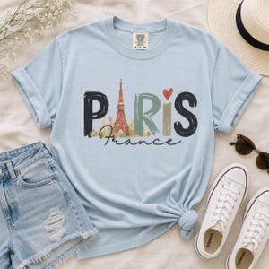 May include: A light blue short-sleeve t-shirt featuring the word PARIS in large, stylized letters. The letter A is replaced by an illustration of the Eiffel Tower, and a small red heart sits above the letter I. The word France is written below.