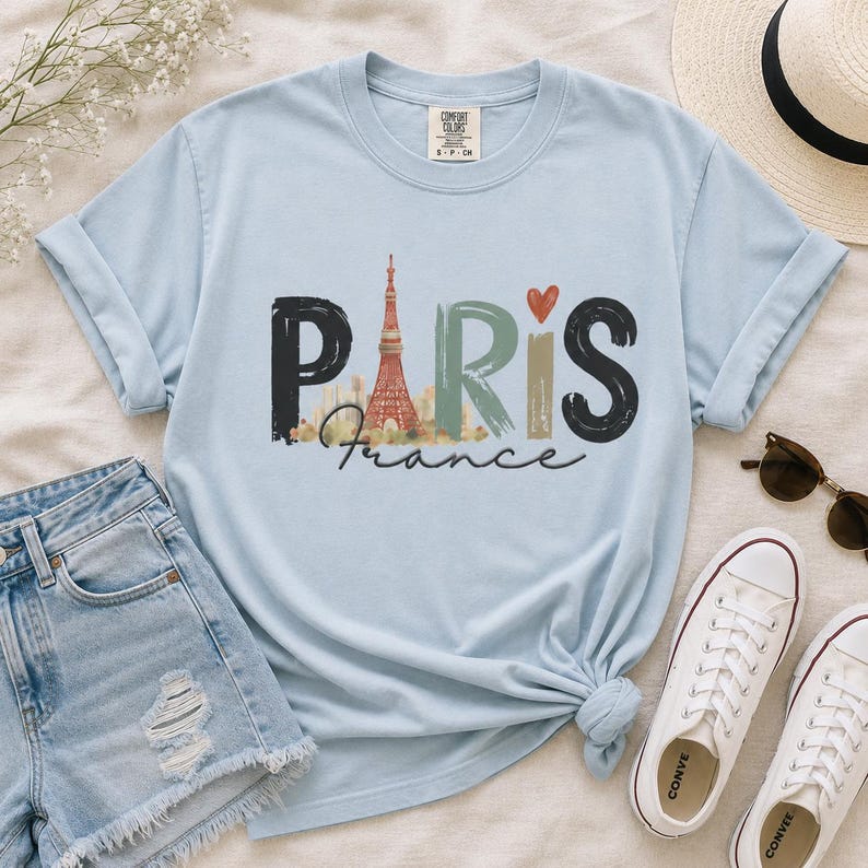 May include: A light blue short-sleeve t-shirt featuring the word PARIS in large, stylized letters. The letter A is replaced by an illustration of the Eiffel Tower, and a small red heart sits above the letter I. The word France is written below.