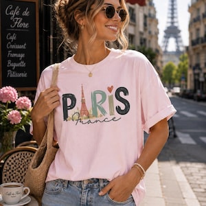 May include: A light pink short-sleeve t-shirt featuring a graphic print with the text PARIS in large, stylized letters, where the letter A is replaced by an illustration of the Eiffel Tower, and the word France is written in cursive below.