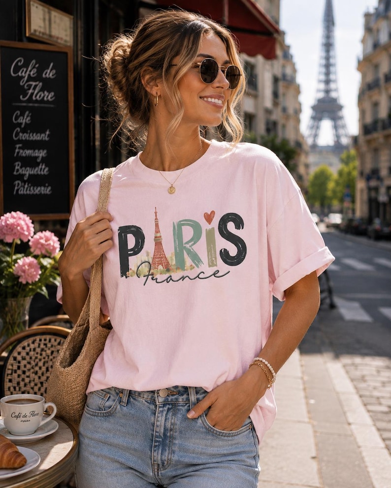 May include: A light pink short-sleeve t-shirt featuring a graphic print with the text PARIS in large, stylized letters, where the letter A is replaced by an illustration of the Eiffel Tower, and the word France is written in cursive below.