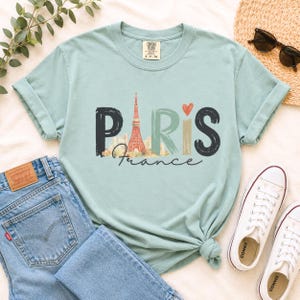 May include: A light sage green short-sleeve t-shirt featuring the word PARIS in large, stylized letters. The letter A is replaced by a graphic of the Eiffel Tower, and a small orange heart sits above the letter I. The word France is written below.