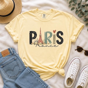 May include: A pale yellow short-sleeve t-shirt featuring the word PARIS in large, stylized letters. The letter A is replaced by a graphic of the Eiffel Tower, and a small red heart sits above the letter I. The word France is written below.