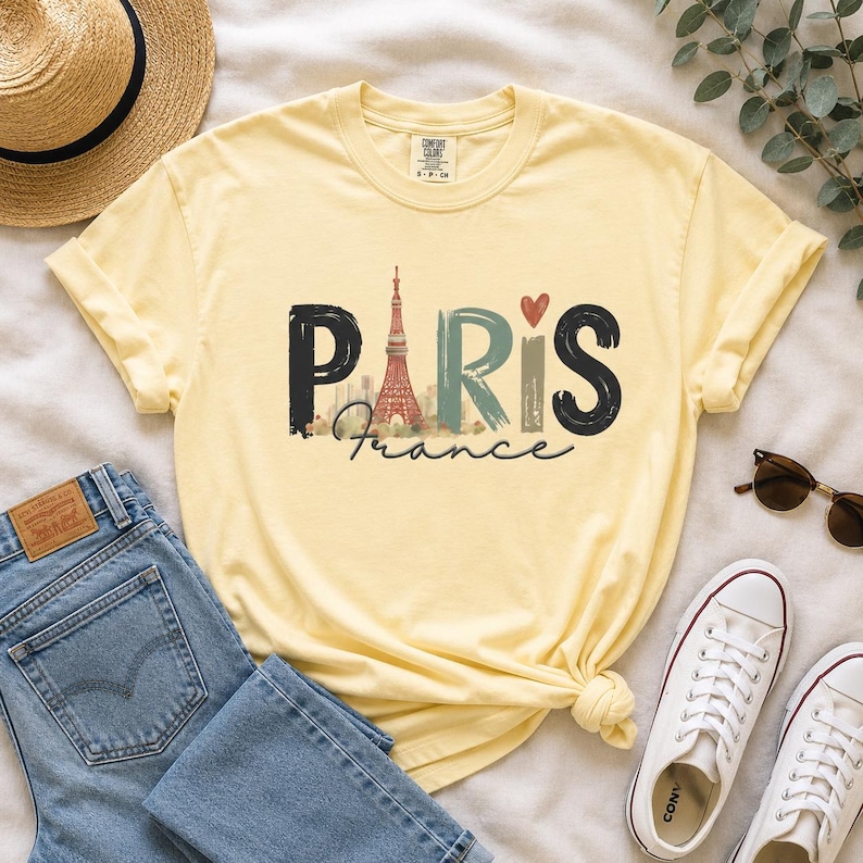 May include: A pale yellow short-sleeve t-shirt featuring the word PARIS in large, stylized letters. The letter A is replaced by a graphic of the Eiffel Tower, and a small red heart sits above the letter I. The word France is written below.