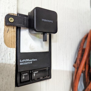 May include: A black LiftMaster Security+ garage door opener with buttons labelled "Lock" and "Light". A black SwitchBot device is mounted above the opener.