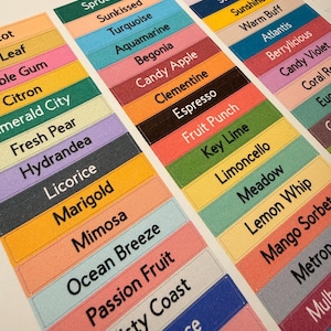 Op de afbeelding: Close-up van kleurrijke rechthoekige labels met verschillende kleurnamen in het zwart gedrukt. De labels zijn in rijen gerangschikt en tonen een reeks tinten van groen en blauw tot roze en oranje. De labels zijn voor een product zoals verf of stof.
