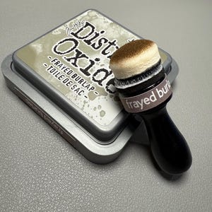 May include: A silver metal ink pad with the text "Distress Oxide - Frayed Burlap - Toile de Sac" and a matching ink blending tool. The ink pad is rectangular and the blending tool has a black handle and a round, layered foam head.