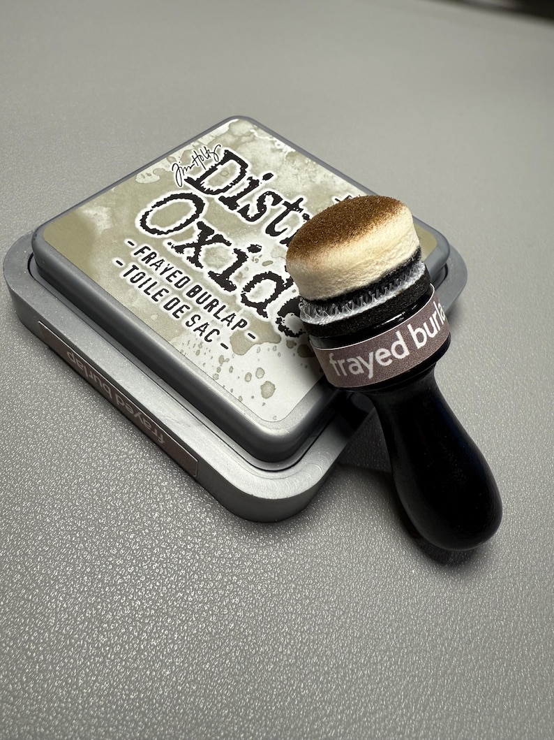 May include: A silver metal ink pad with the text "Distress Oxide - Frayed Burlap - Toile de Sac" and a matching ink blending tool. The ink pad is rectangular and the blending tool has a black handle and a round, layered foam head.