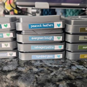 May include: A stack of gray ink pads with labels that read "peacock feathers", "evergreen bough", "salvaged patina", and "tumbled glass". Each label has a small heart icon. The ink pads are on a textured surface.