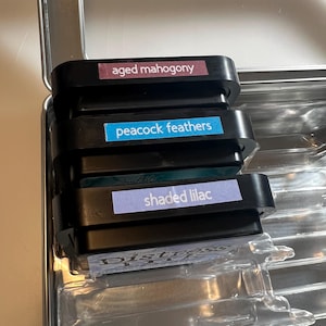 May include: A stack of three black ink pads with labels reading "aged mahogany", "peacock feathers", and "shaded lilac". The ink pads are inside a silver metal case with clear plastic inserts.