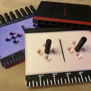 May include: Three 3D-printed tools with black and colored accents. One is lavender with the text "Moms Mums & Memes" and a ruler along the bottom. Another is black with red accents. The third is pink with black ruler markings.