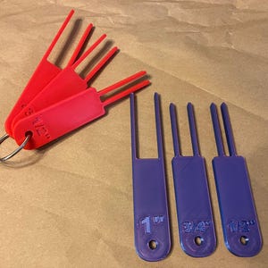 May include: Red and purple plastic tools with measurement markings. The red tools are attached to a metal ring. The purple tools are marked with 1", 3/4", and 1/2" measurements. The tools have a handle with prongs.