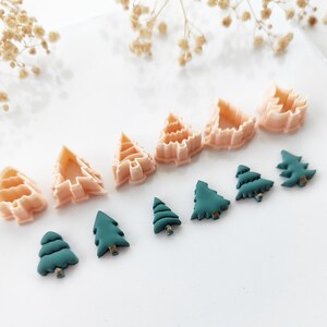 Può includere: Una collezione di formine per biscotti e ornamenti a forma di albero di Natale. Le formine sono color pesca chiaro e gli ornamenti sono verde acqua. Gli oggetti sono disposti su una superficie bianca, con fiori secchi sullo sfondo.
