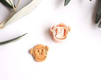 May include: A monkey-shaped cookie cutter and a baked cookie are displayed on a white surface, with a sprig of green leaves in the background. The cookie cutter is light pink, and the cookie is tan.