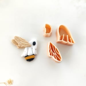 Puede incluir: Un conjunto de cortadores de galletas con forma de abeja en naranja claro y blanco. La abeja tiene alas de color canela, un cuerpo blanco y un abdomen a rayas negras y amarillas. También hay tres piezas separadas de cortadores de galletas en naranja claro.