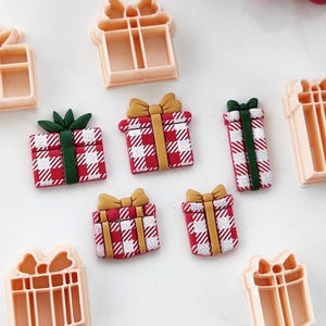 Könnte beinhalten: Eine Sammlung von weihnachtlichen Ausstechformen und Geschenk-förmigen Dekorationen. Die Ausstecher sind aus hellpfirsichfarbenem Kunststoff, während die Ornamente rot-weiß karierte Muster mit grünen oder goldenen Schleifen und Bändern aufweisen. Festliche Weihnachtsdekoration.