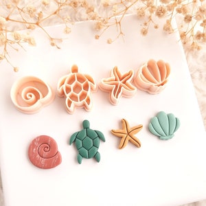 Może przedstawiać: Zestaw foremek do ciastek o tematyce morskiej, w tym muszla, żółw, rozgwiazda i muszla przegrzebka. Foremki są w odcieniach beżu, zieleni i koralu, ułożone na białej powierzchni. Dekoracyjne gałązki suszonych kwiatów w tle.