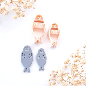 Peut inclure: Ensemble de découpoirs à biscuits et moules à fondant en forme de phoque. Deux découpoirs en plastique rose et deux moules à fondant gris sont disposés sur une surface blanche. Les découpoirs et moules sont de tailles différentes.